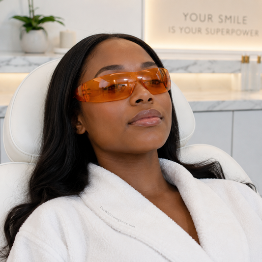 A black woman in her late 20s reclines in a white spa treatment chair wearing orange wraparound protective teeth whitening glasses. The modern whitening studio features white cabinetry, marble-style counters, warm under-cabinet lighting, a potted plant, and a wall sign reading “Your Smile Is Your Superpower.”